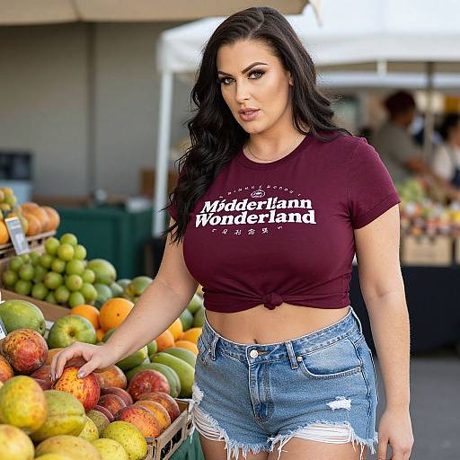 Photograph of a curvy woman with long black hair, wearing a tied maroon crop top and distressed denim shorts, standing in a colorful fruit market