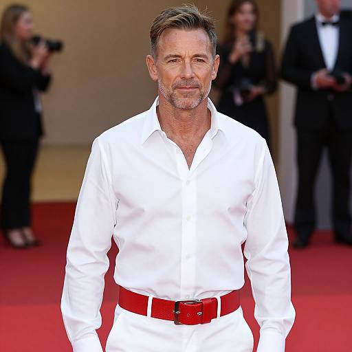 Photograph of mature, handsome man with gray hair and beard, wearing white shirt, red belt, and white pants, standing on red carpet. Background