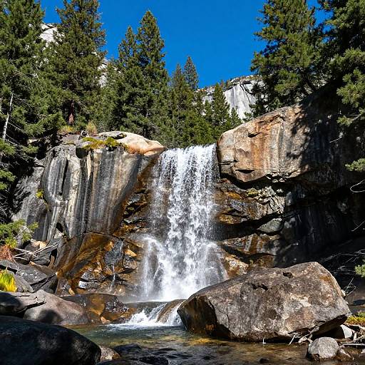 Photograph of a cascading waterfall in a rocky, forested mountain landscape with tall evergreen trees and clear blue sky.