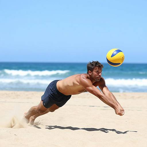 Photograph of a muscular, tan-skinned man in black swim trunks, diving to hit a yellow and blue volleyball on a sunny, sandy beach