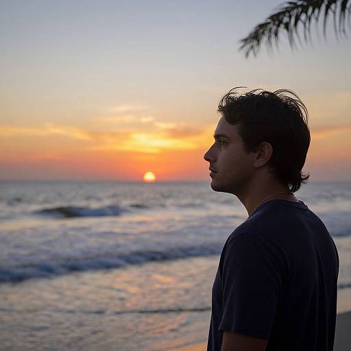 Photograph of a young man in silhouette, profile view, standing on a beach at sunset with orange and blue sky, ocean waves, and palm fr