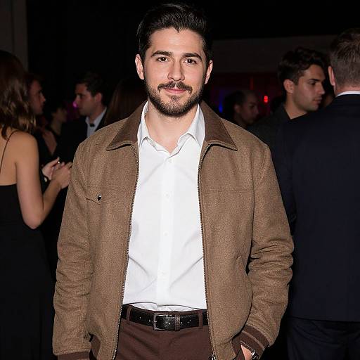 Photograph of a bearded man with dark hair, wearing a brown jacket over a white shirt, standing in a dimly lit, crowded social event