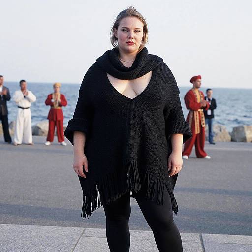 Photograph of a plus-size woman with fair skin, brown hair in a ponytail, wearing a black fringe shawl and black pants, standing on