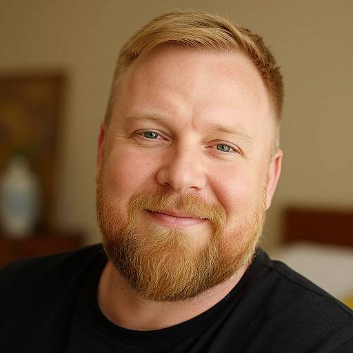 Photograph of a smiling, fair-skinned, bearded man with red hair, blue eyes, wearing a black shirt, in a softly lit bedroom