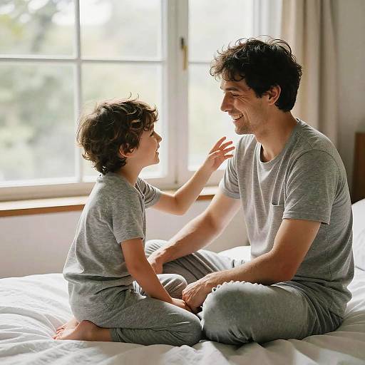 Photograph of smiling father and curly-haired son in matching gray pajamas, sitting on a sunlit bed, laughing together.