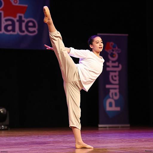 Photograph of a young Asian woman performing a high kick on stage, wearing a white shirt and beige pants, with 