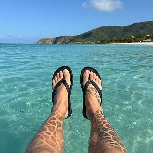 Photograph of tattooed legs wearing black flip-flops in clear turquoise water, with a mountainous coastline and blue sky in the background.