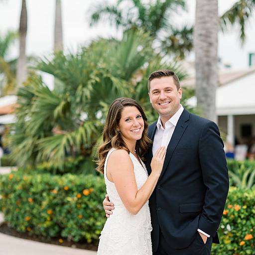 Photograph of a smiling couple; the woman in a white lace dress, the man in a black suit, standing outdoors with palm trees and greenery