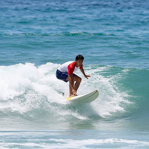 Photograph of a young male surfer in a red-and-white shirt and blue shorts riding a small wave in vibrant blue ocean water.