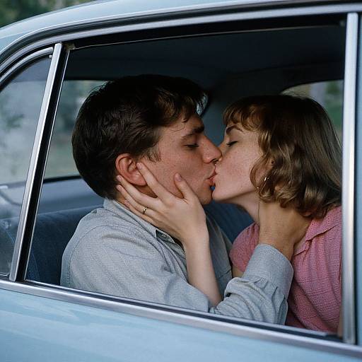 Photograph of a young couple passionately kissing inside a silver vintage car, with the woman gently holding the man's face.