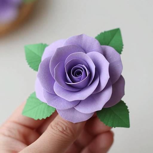 Photograph of a delicate, paper-crafted lavender rose with green leaves, held by a hand against a softly blurred white background.