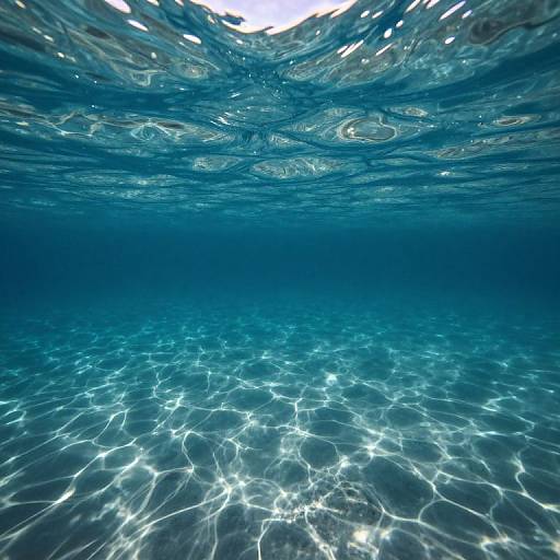 Underwater photograph showing clear blue water with sunlight creating intricate, wavy patterns on the sandy ocean floor.