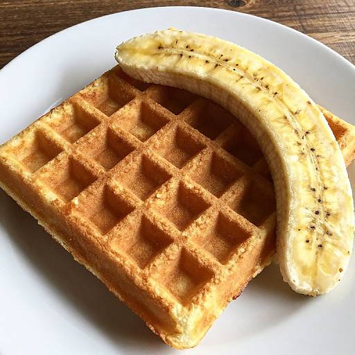 Photograph of a golden-brown waffle with a slightly charred texture, topped with a curved, peppered banana slice on a white plate.