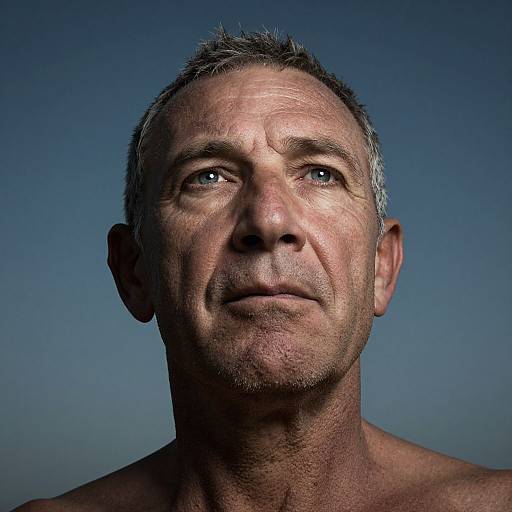 Photograph of a middle-aged, shirtless man with short gray hair, blue eyes, and weathered skin, against a clear blue sky background.
