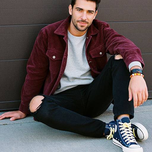 Photograph of a bearded man with short dark hair, wearing a maroon corduroy jacket, white t-shirt, black ripped jeans, and