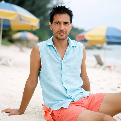 Photograph of a smiling, tan-skinned man with short dark hair, wearing a sleeveless light blue shirt and bright orange shorts, sitting on a