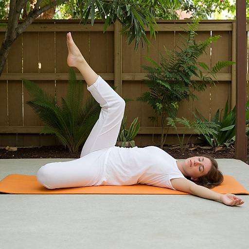 Photograph of a woman in white yoga clothes lying on an orange mat, performing a leg stretch outdoors with greenery and wooden fence in the background.
