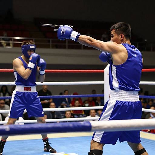 Photograph of two male boxers in blue and white uniforms, one throwing a punch, the other defending, in a brightly lit boxing ring.