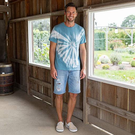 Photograph of a smiling man with short brown hair, wearing a blue tie-dye t-shirt, denim shorts, and white slip-on shoes, standing