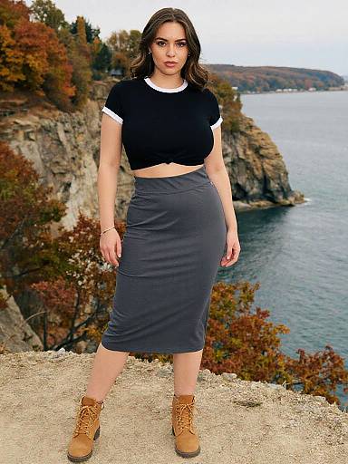 Photograph of a curvy woman with dark hair, wearing a black crop top, gray skirt, and tan boots, standing on a rocky cliff overlooking