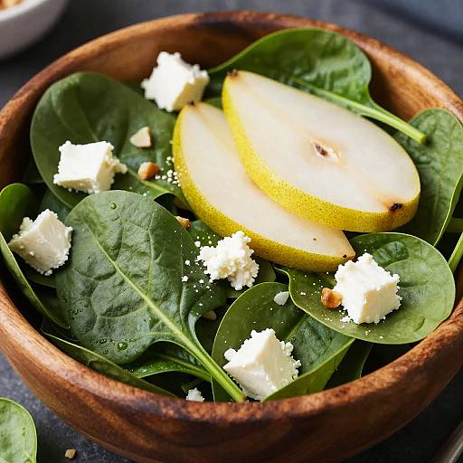 Photograph of a wooden bowl filled with fresh spinach leaves, crumbled feta cheese, and sliced yellow pear, with small red pepper bits scattered on