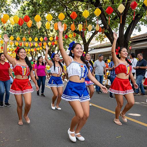 Photograph of five smiling Latina women in colorful Mexican-themed costumes, dancing on a street with yellow and red paper lanterns, surrounded by cheering spectators and