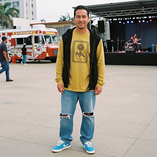 Photograph of a smiling man with short black hair, wearing a yellow shirt, black vest, ripped jeans, and blue sneakers, standing in an outdoor
