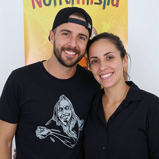 Photograph of a smiling, bearded man with a black cap and graphic t-shirt, standing close to a smiling woman with black hair in a pony