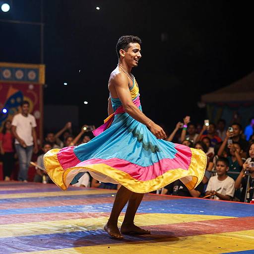 Photograph of a male dancer in a colorful, sleeveless, blue dress with pink and yellow trim, performing on a brightly lit circus stage with an