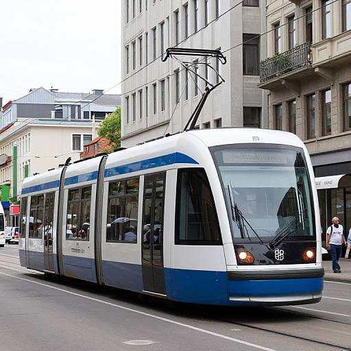 Photograph of a modern, white and blue streetcar with black roof poles, traveling on a city street lined with tall buildings.
