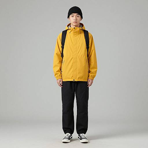 Photograph of an Asian teenage boy in a yellow hooded jacket, black beanie, black pants, white sneakers, and backpack, standing against a