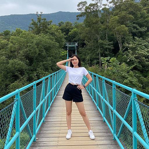 Photograph of a young woman with fair skin, black shorts, white t-shirt, and white sneakers, standing confidently on a turquoise bridge amidst lush green