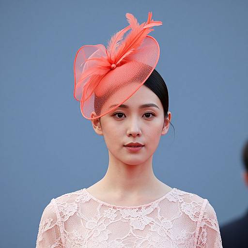 Photograph of an Asian woman with fair skin, wearing a white lace dress and a vibrant red feathered fascinator, set against a plain blue background