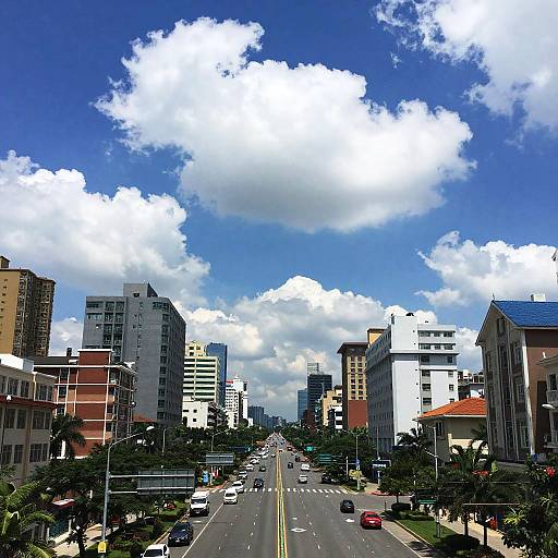 Photograph of a modern urban street with tall buildings, blue sky, and fluffy white clouds; cars and trees line the road.