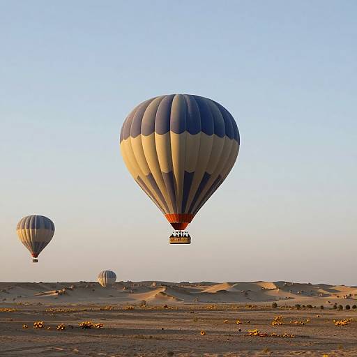 Photograph of two colorful hot air balloons soaring over a sandy desert with rolling dunes at sunrise, with another smaller balloon in the background.