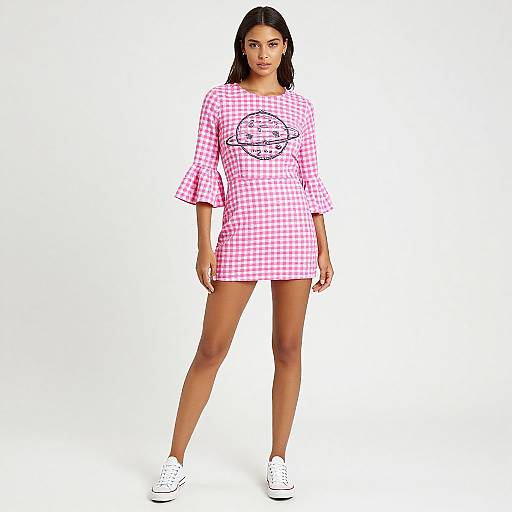 Photograph of a young woman with dark hair, wearing a pink and white gingham dress with a black graphic, and white sneakers, standing against a