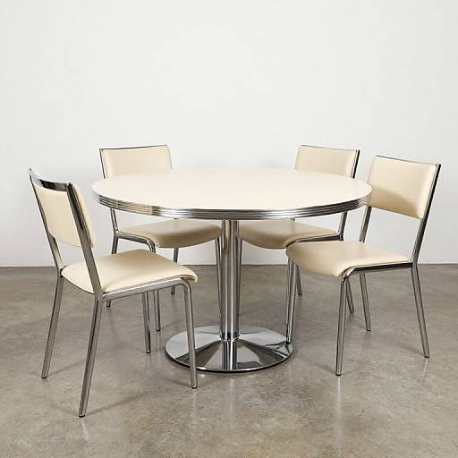 Photograph of a modern, round, chrome-finished table with four beige leather chairs, set on a gray concrete floor against a white wall.