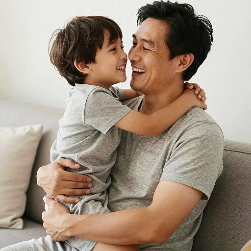 Photograph of an Asian man in a gray t-shirt, smiling while hugging a young boy in a matching gray shirt, seated on a gray sofa