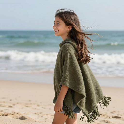 Young girl with long brown hair, wearing an olive-green fringed shawl and denim shorts, smiling on a sandy beach with gentle waves in the background
