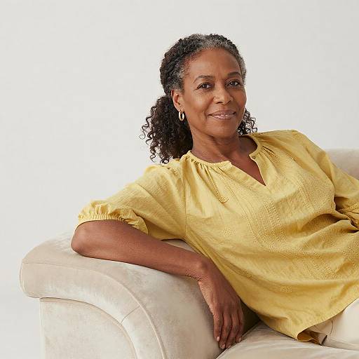 Photograph of a smiling African-American woman with curly hair, wearing a yellow blouse, resting on a white couch against a plain white background.
