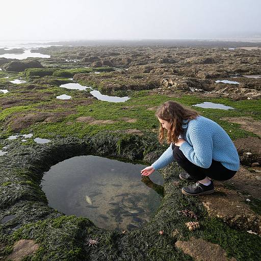 Photograph of a woman with long brown hair, wearing a blue sweater and black pants, crouching by a small, reflective pond in a rocky