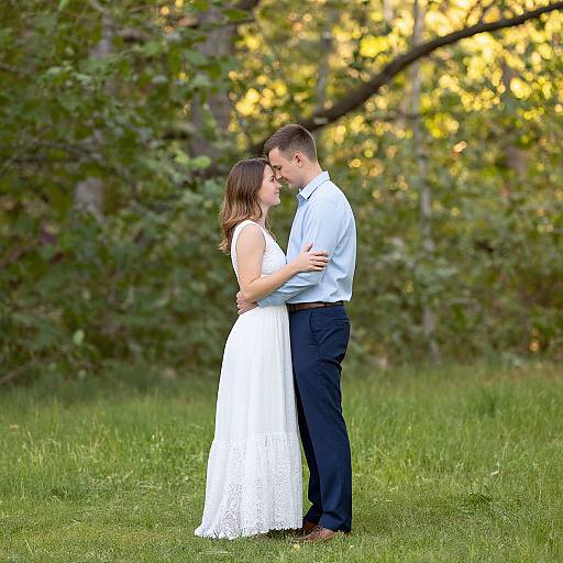Photograph of a couple standing in a grassy field, both gazing into each other's eyes. The woman wears a white lace dress, the
