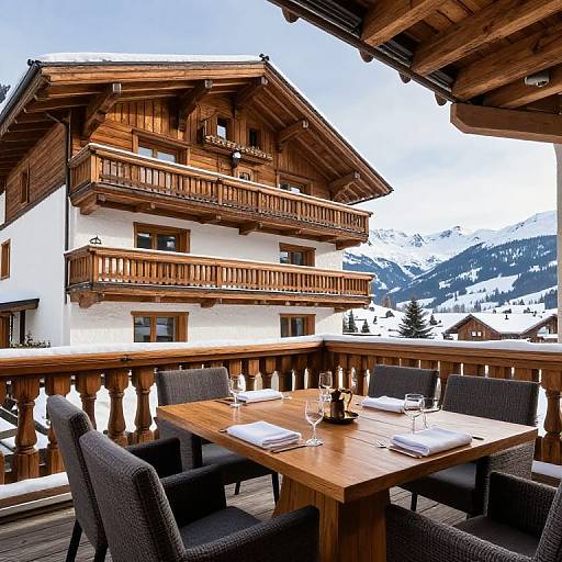 Photograph of a rustic wooden chalet with balconies, set on a snowy mountain terrace, featuring a wooden dining table with black chairs and glassware
