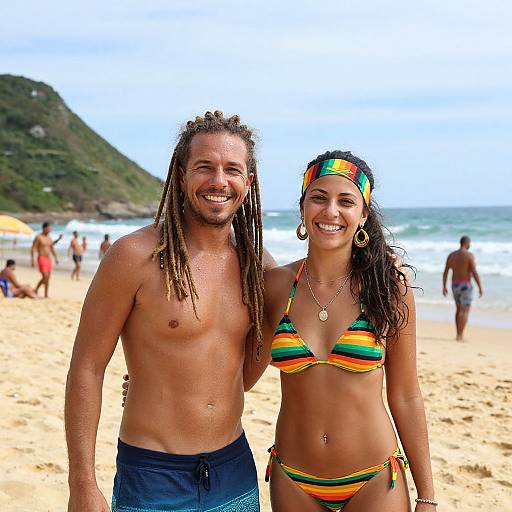 Photograph of a smiling, shirtless man with dreadlocks and a woman in a colorful striped bikini, standing on a sandy beach with ocean waves and