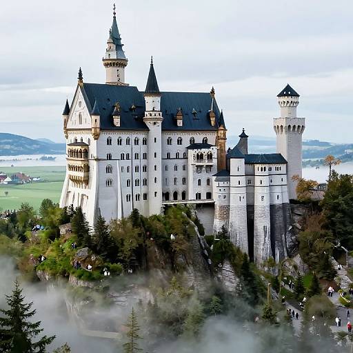 Photograph of a grand, white, medieval-style castle with multiple black-topped towers, surrounded by lush green trees and mist, set against a bright