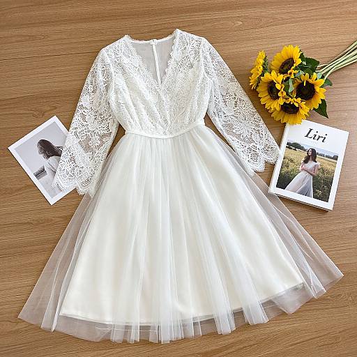 Photograph of a white lace long-sleeve dress with a sheer tulle skirt, laid on a wooden table with sunflowers, a photo of