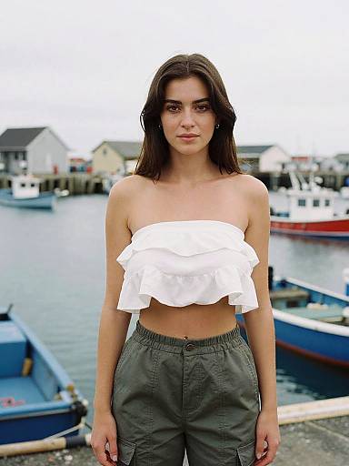 Photograph of a young woman with straight brown hair, wearing a white ruffled tube top and olive green pants, standing in front of a marina