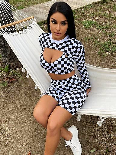 Photograph of a curvy woman with long black hair, wearing a black-and-white checkered crop top and skirt, sitting on a white hammock