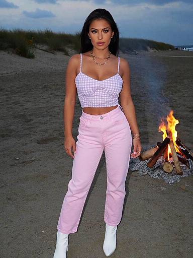 Photograph of a dark-haired woman with medium skin tone, wearing a checkered crop top, pink pants, and white boots, standing in front of