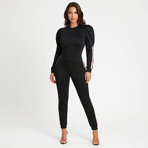 Photograph of a confident woman with wavy dark hair, wearing a black long-sleeve top and matching black pants with red stripes, standing against